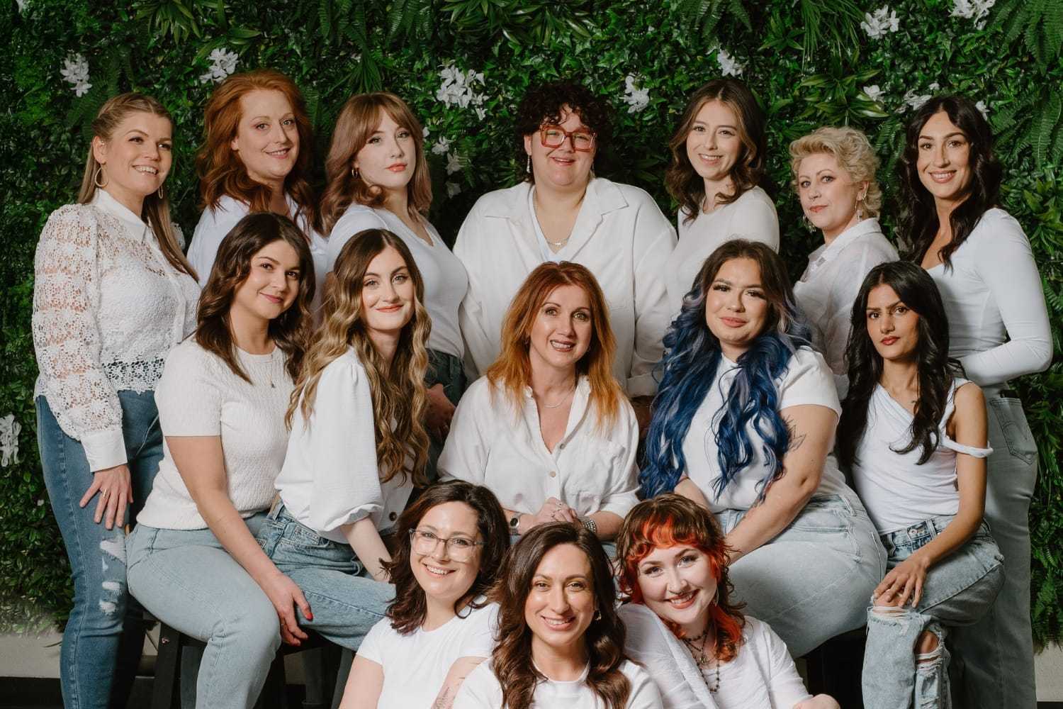 Group of women in white tops and jeans posing in front of green foliage backdrop.